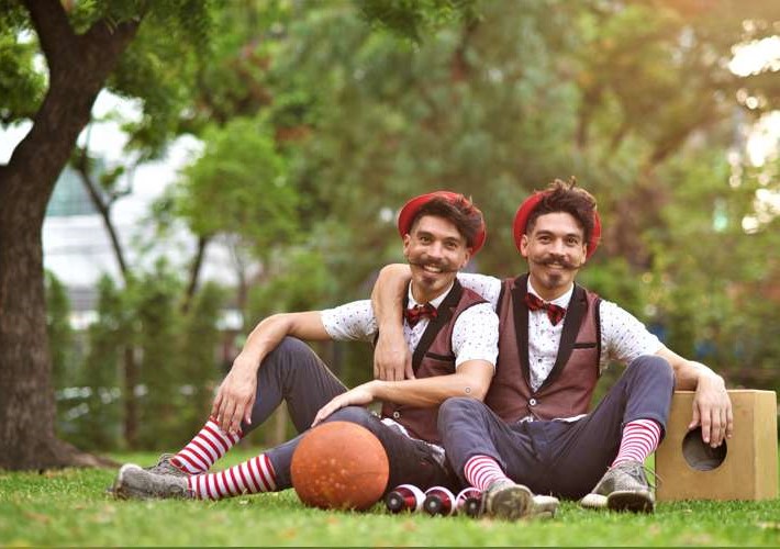 Two men sitting on the grass. They wear the same clothes. In front of them there is a basketball and juggling clubs